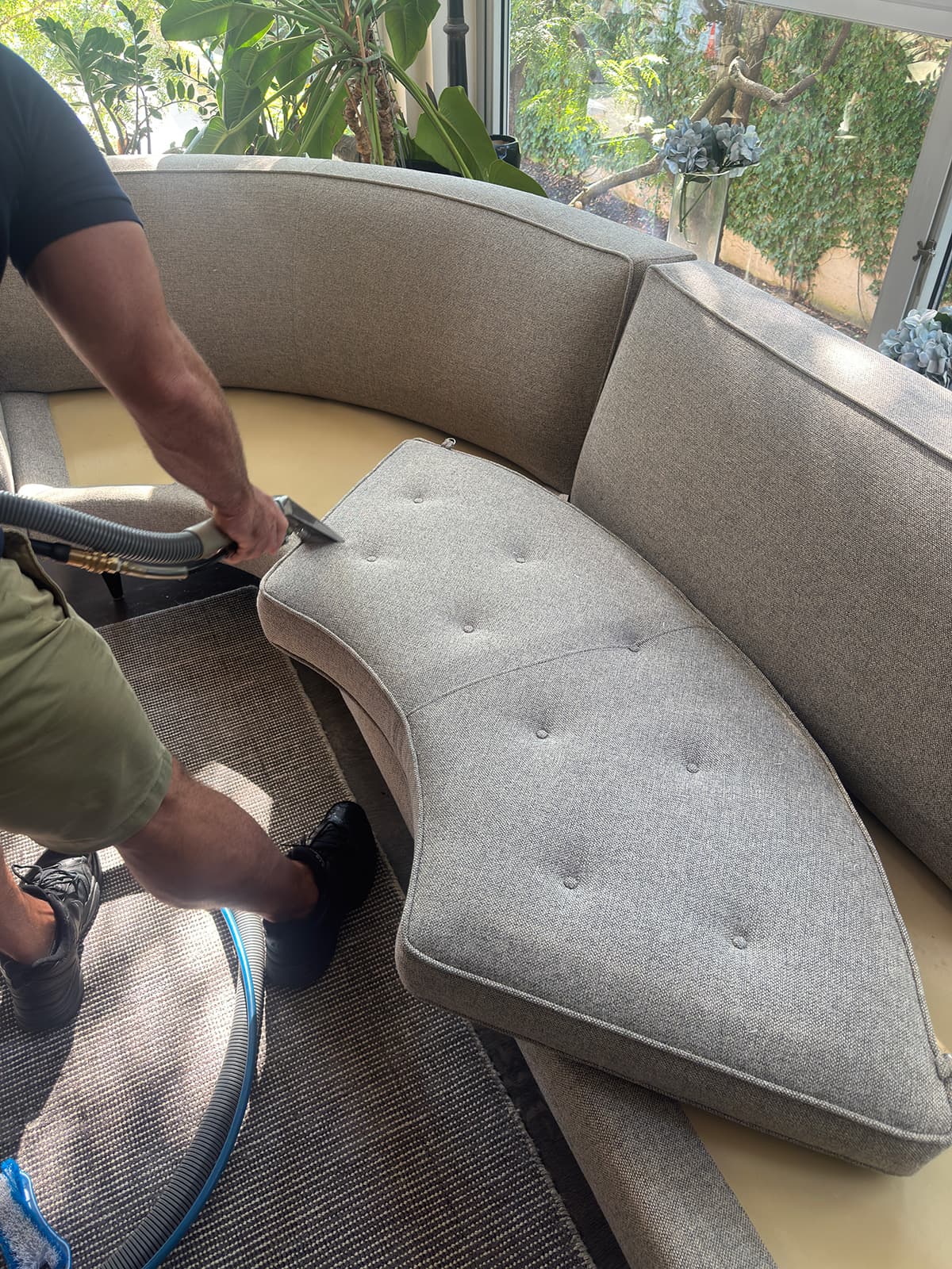 A technician running an upholstery tool along the seat of a curved grey tufted sofa